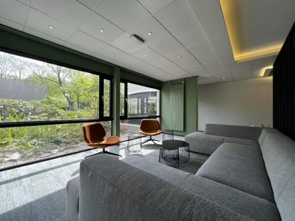 Moderne wachtruimte met grijze hoekbank, twee oranje stoelen en grote ramen met uitzicht op een groene tuin bij Verderlaan.